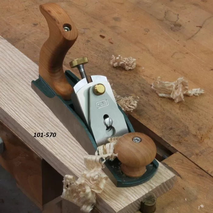 Bench hand plane smoothing wood board on workbench, labeled 101-570, with wood shavings.