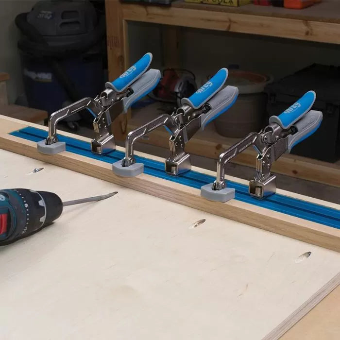 Four toggle clamps with blue handles mounted on a T-track, woodworking drill nearby