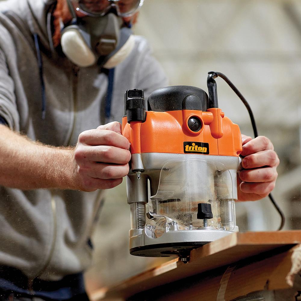 Woodworker using an orange Triton plunge router on a wooden board in a workshop