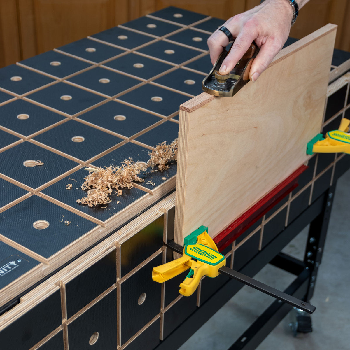 Woodworker planing plywood edge on grid-patterned workbench with clamps and wood shavings