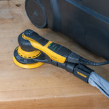 A yellow and black Mirka orbital sander with a gray dust hose attached, resting on a light-colored wooden workbench next to a black dust collector.