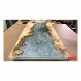 Pouring clear epoxy resin into a live edge wood slab river table on a workshop bench