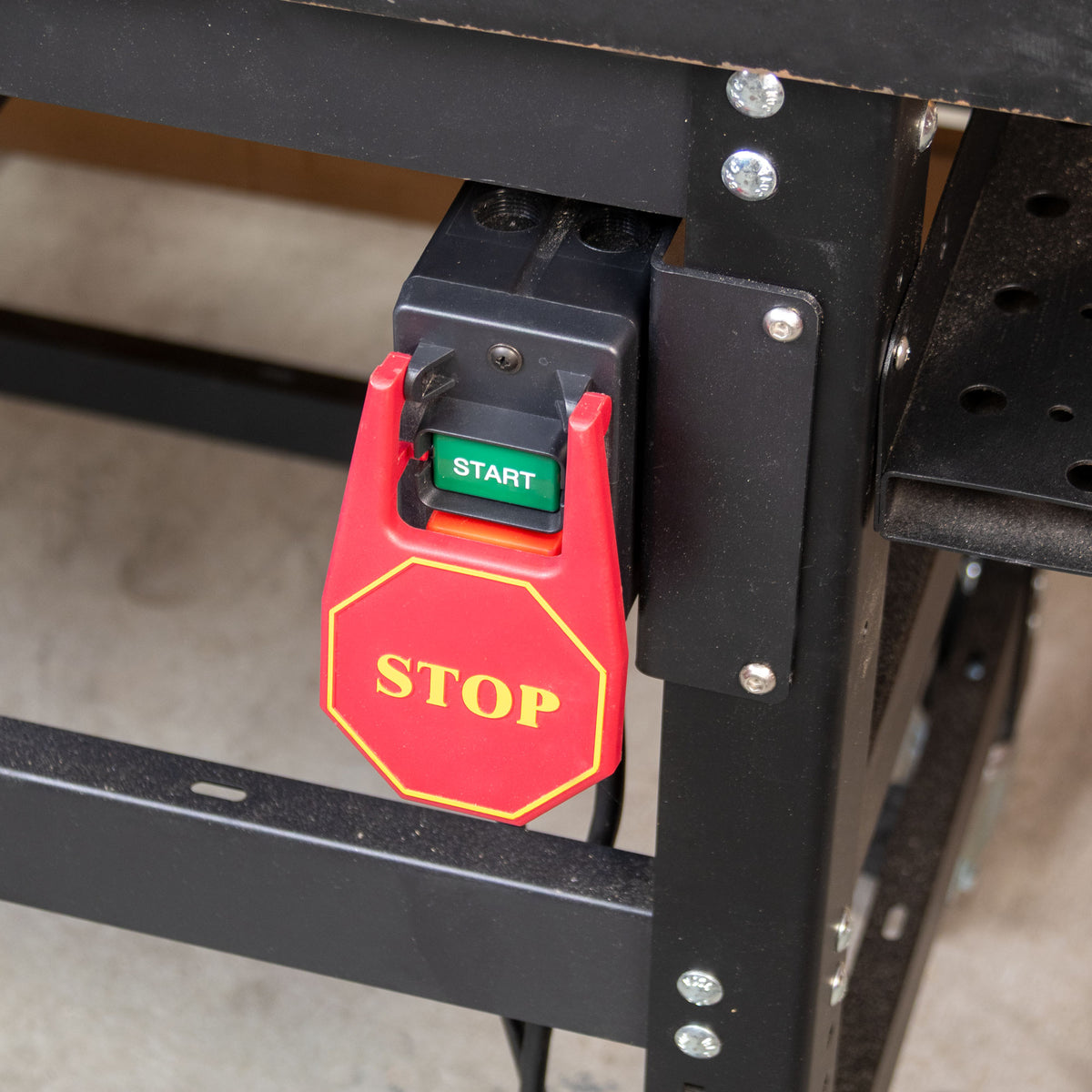A red and black safety stop/start switch mounted on a black metal workbench frame, with a red octagonal stop sign design and a green start button.
