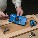 Person using a blue Kreg cabinet hardware jig on a wooden cabinet door with hinges and a router bit nearby.