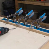 Four toggle clamps with blue handles mounted on a T-track, woodworking drill nearby