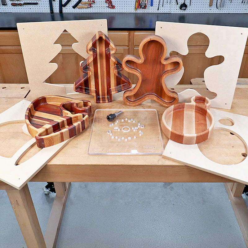 Striped wooden holiday trays shaped like a tree, gingerbread figure, and ornament with woodworking router jigs on a workbench.
