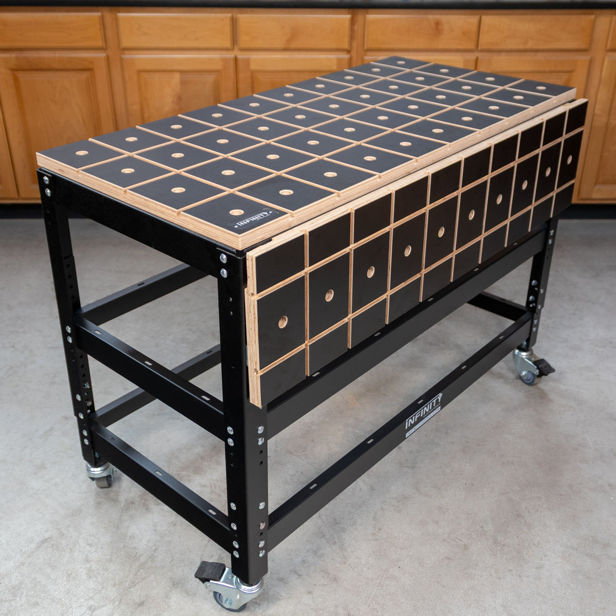 Mobile woodworking workbench with black grid-patterned top, durable steel frame, and caster wheels in a workshop setting.