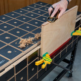 Woodworker planing plywood edge on grid-patterned workbench with clamps and wood shavings