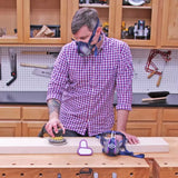 Woodworker sanding board with orbital sander in workshop, wearing protective mask