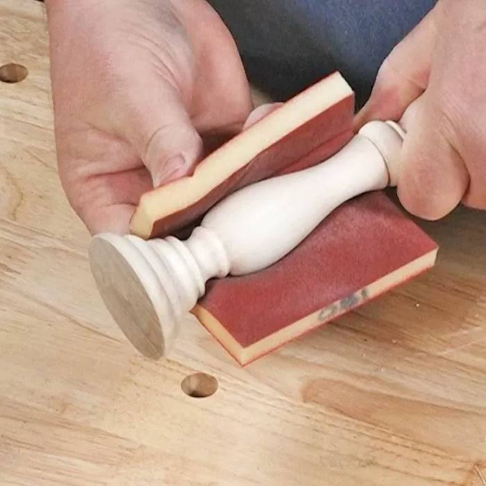 Hand sanding a turned wood spindle with foam sanding blocks on a workbench